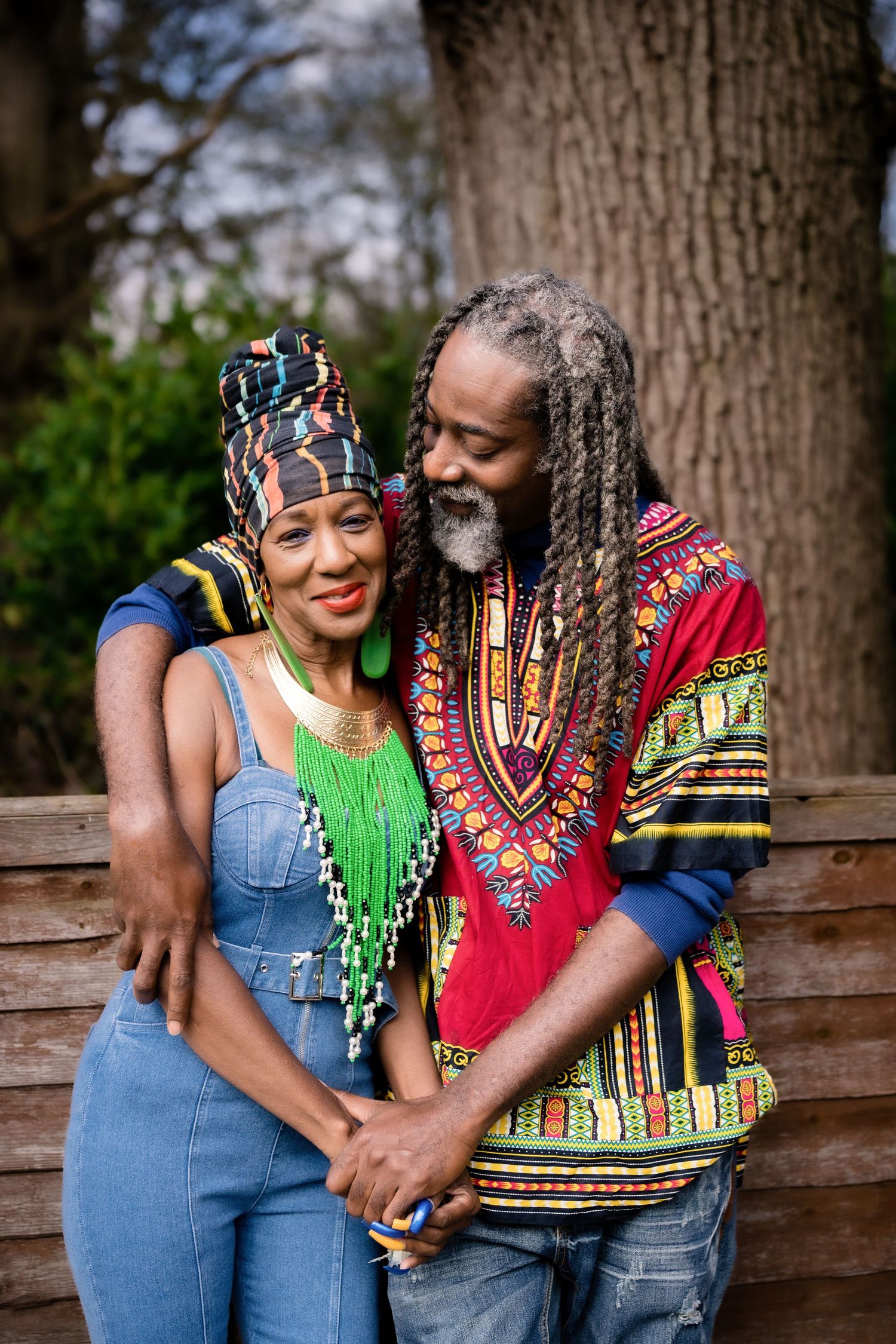 A couple wearing colorful African-inspired clothing embrace while standing against a wooden fence outdoors.