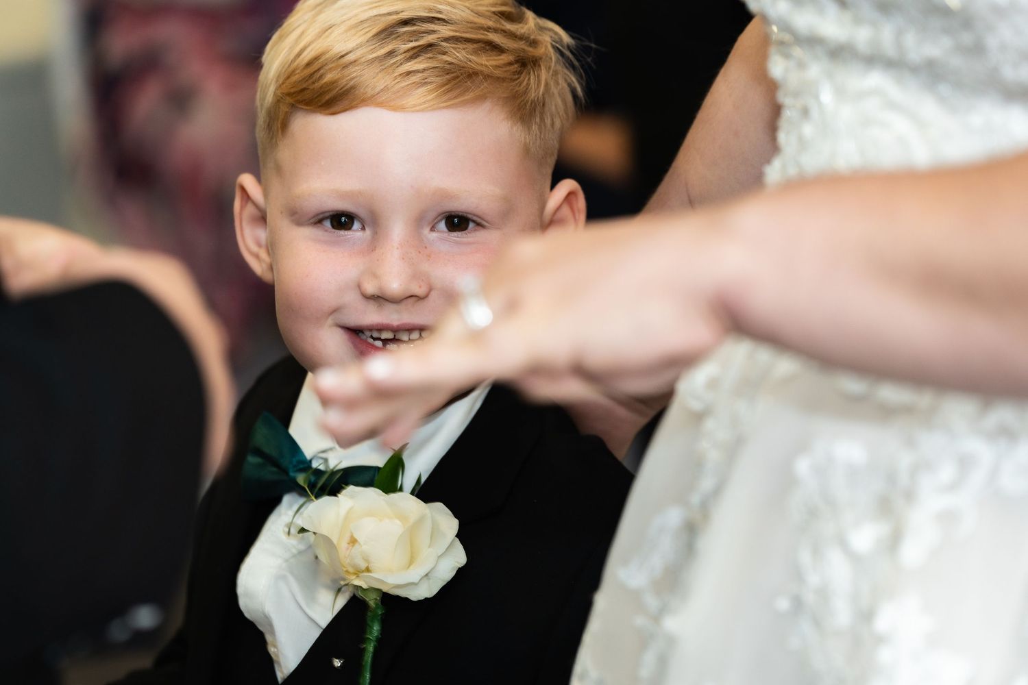 Young ring bearer in black tuxedo smiles during wedding ceremony.