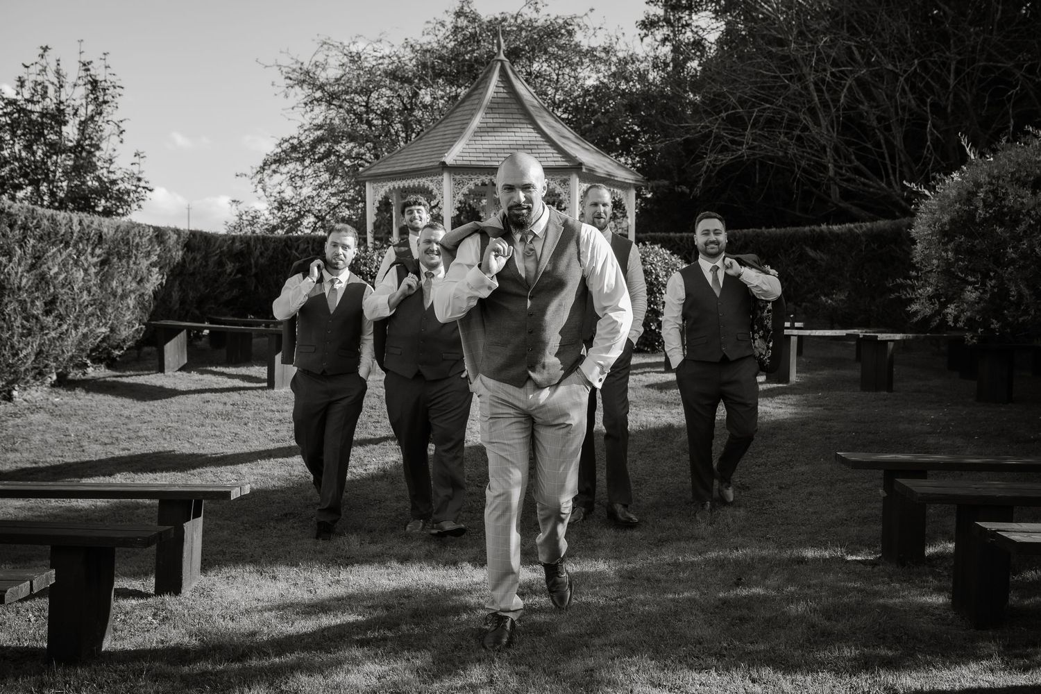 Groom and groomsmen running joyfully down outdoor aisle in black and white photograph.