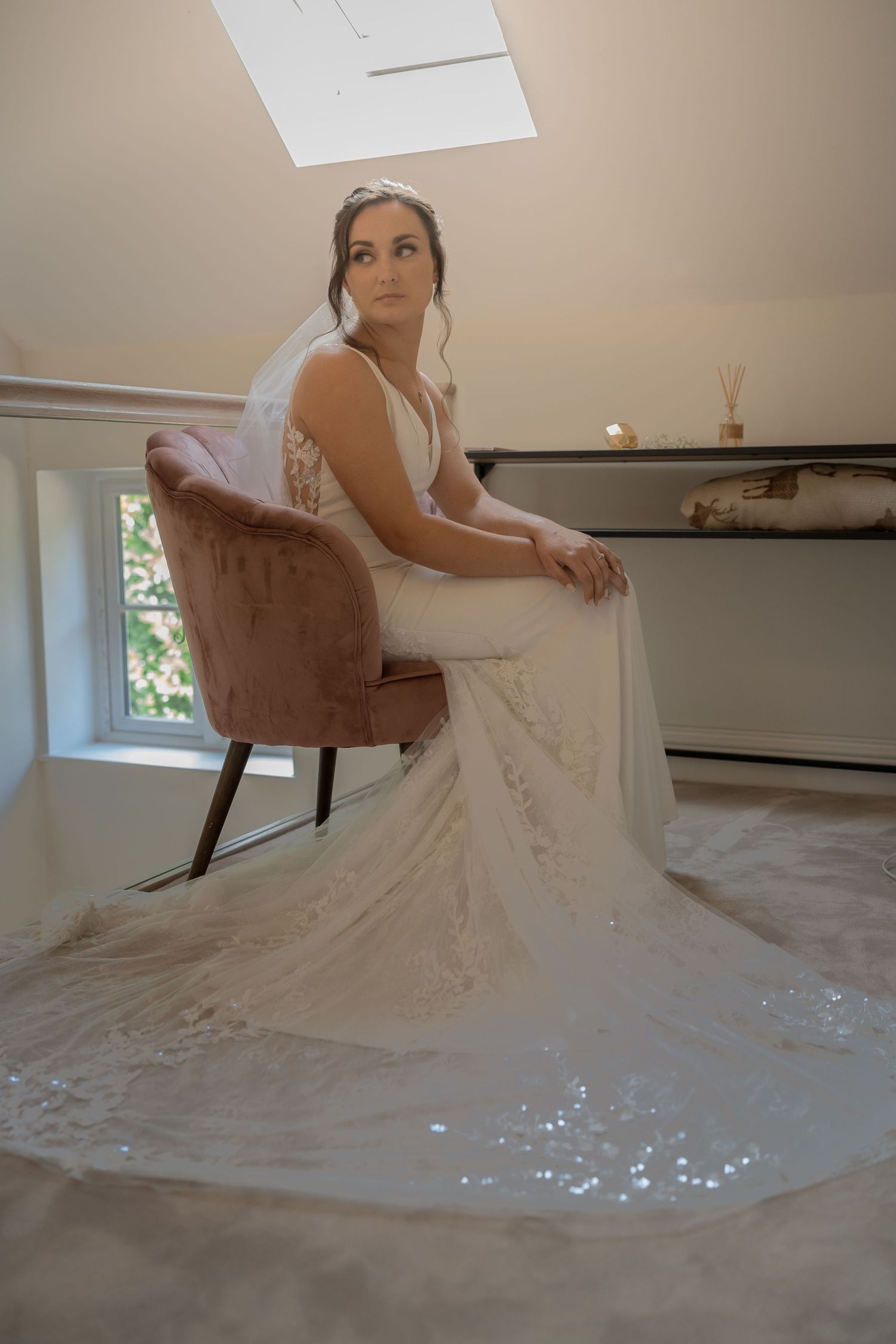 A white lace wedding dress with an elegant train flows around a velvet chair in a sunlit room.