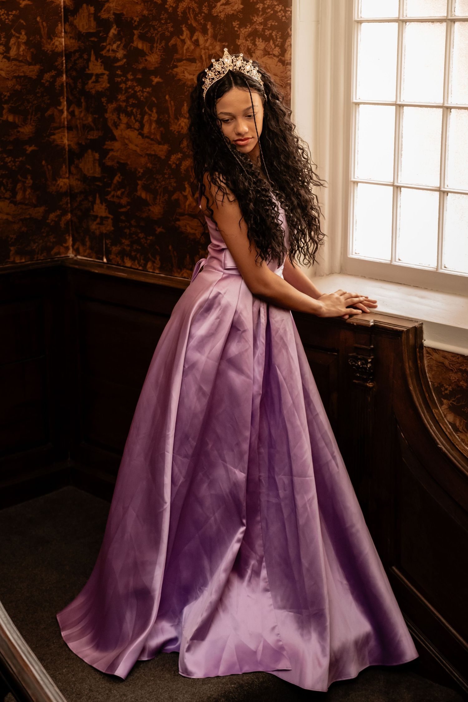 A person wearing a crown and purple ball gown poses gracefully in an elegant interior setting.