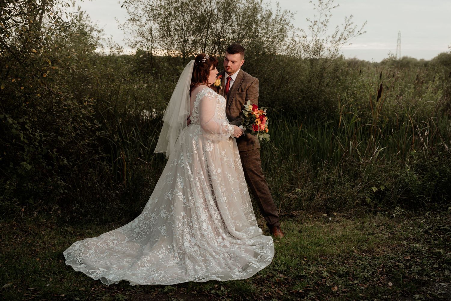 A couple shares an intimate moment in their wedding attire against a natural outdoor backdrop at sunset.