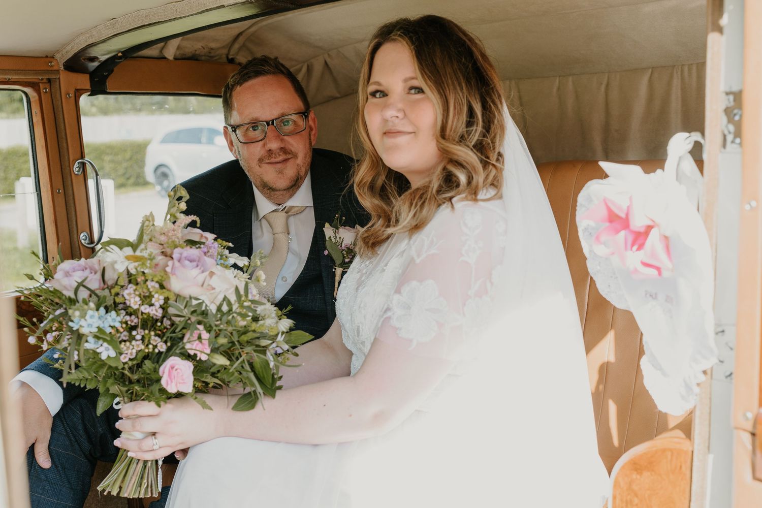 A wedding couple sits together in a vintage vehicle with fresh floral bouquet.