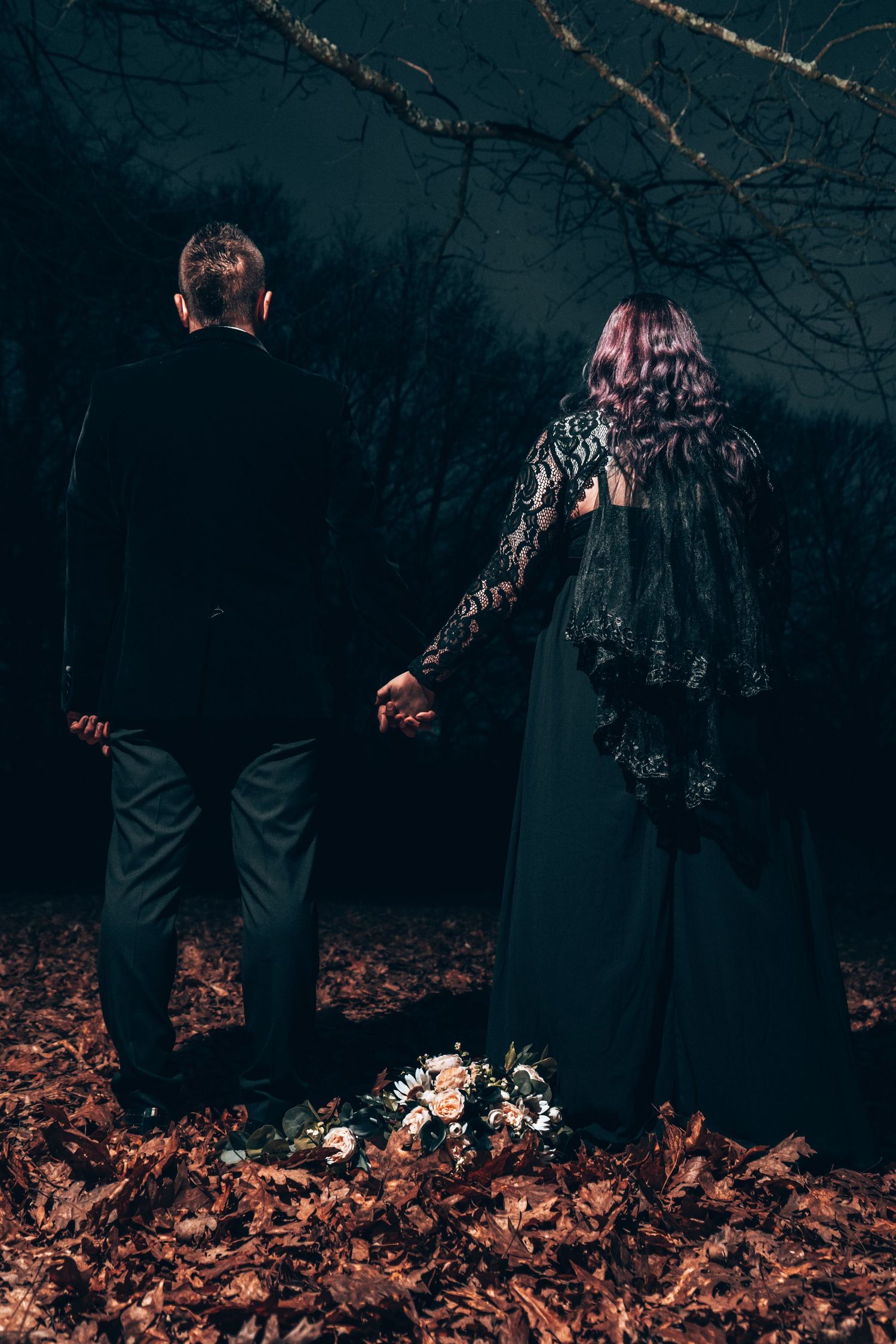 Moody engagement photo of a couple holding hands among autumn leaves at dusk.