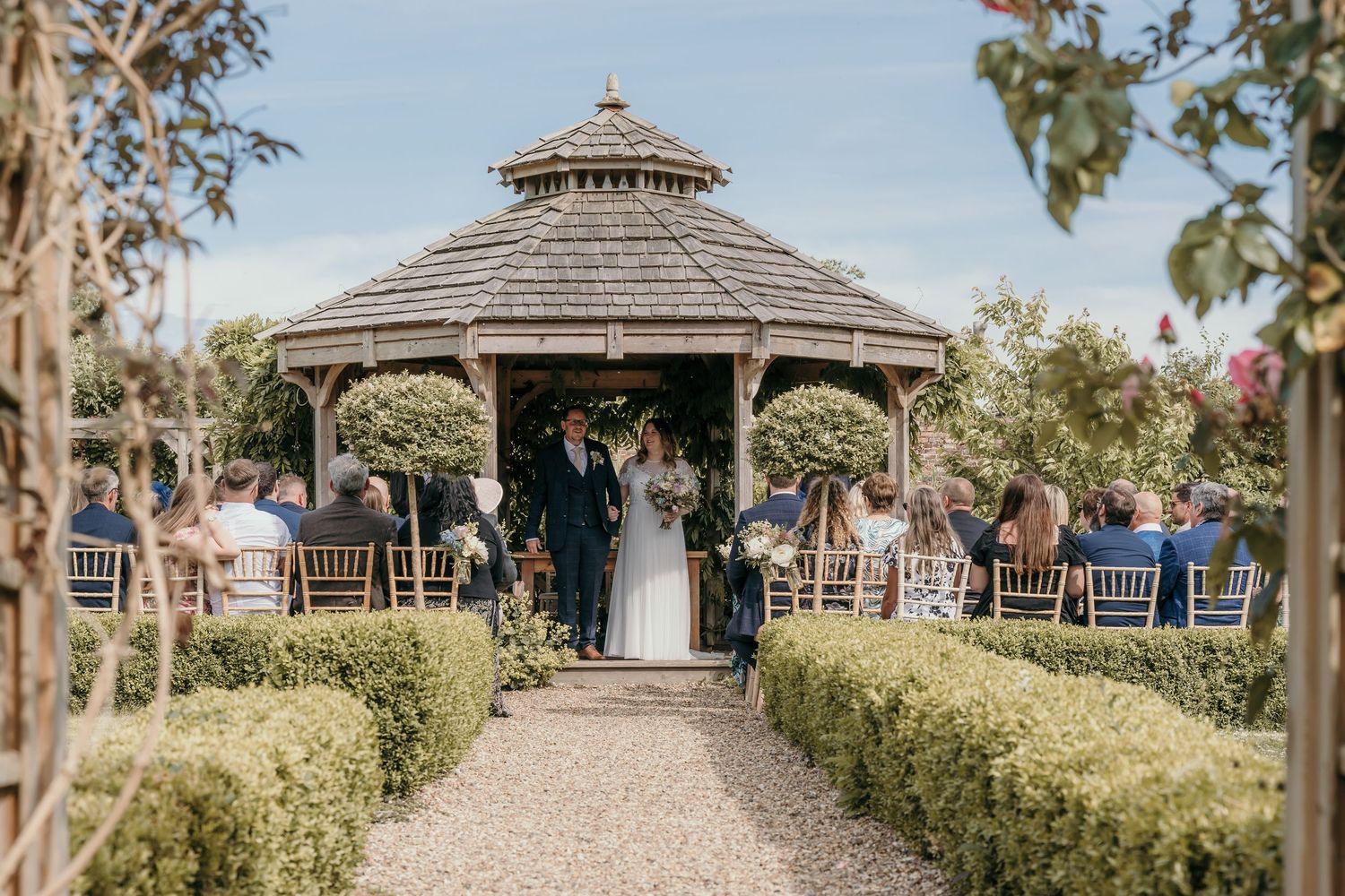 A garden wedding ceremony takes place in an ornate wooden gazebo surrounded by manicured hedges.