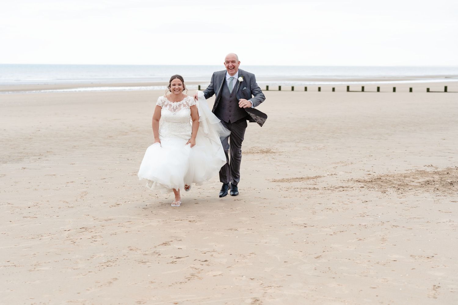 A joyful couple runs along the beach with waves and groynes in the background.