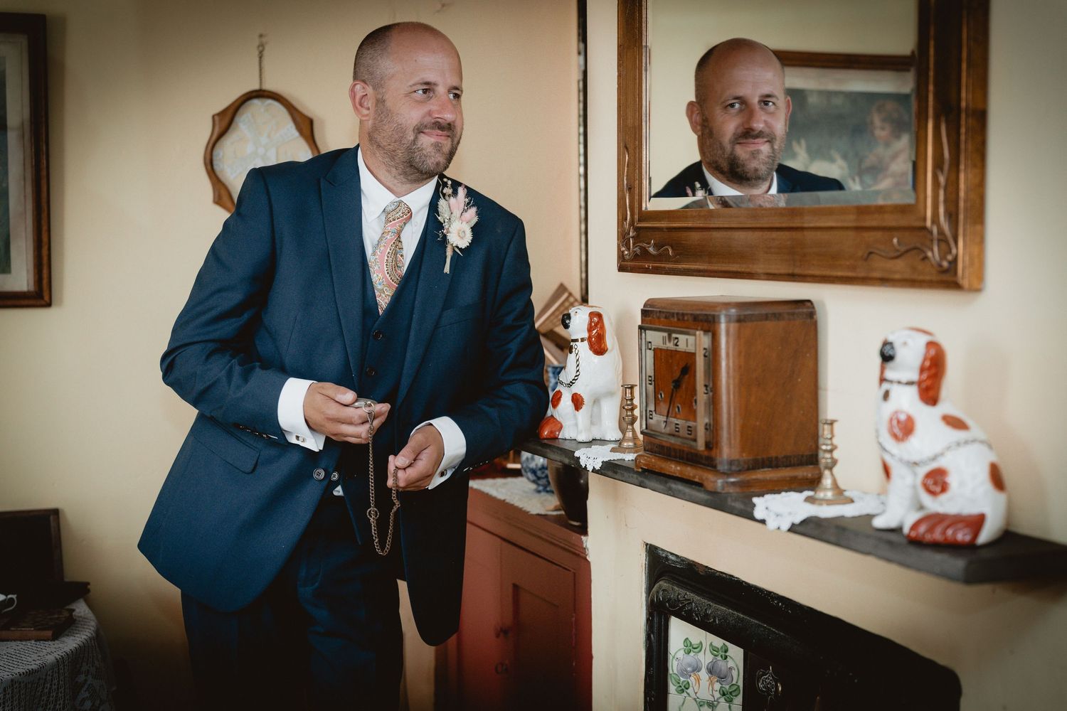 A person in a navy blue suit adjusts their attire while standing next to a mirror and decorative items.