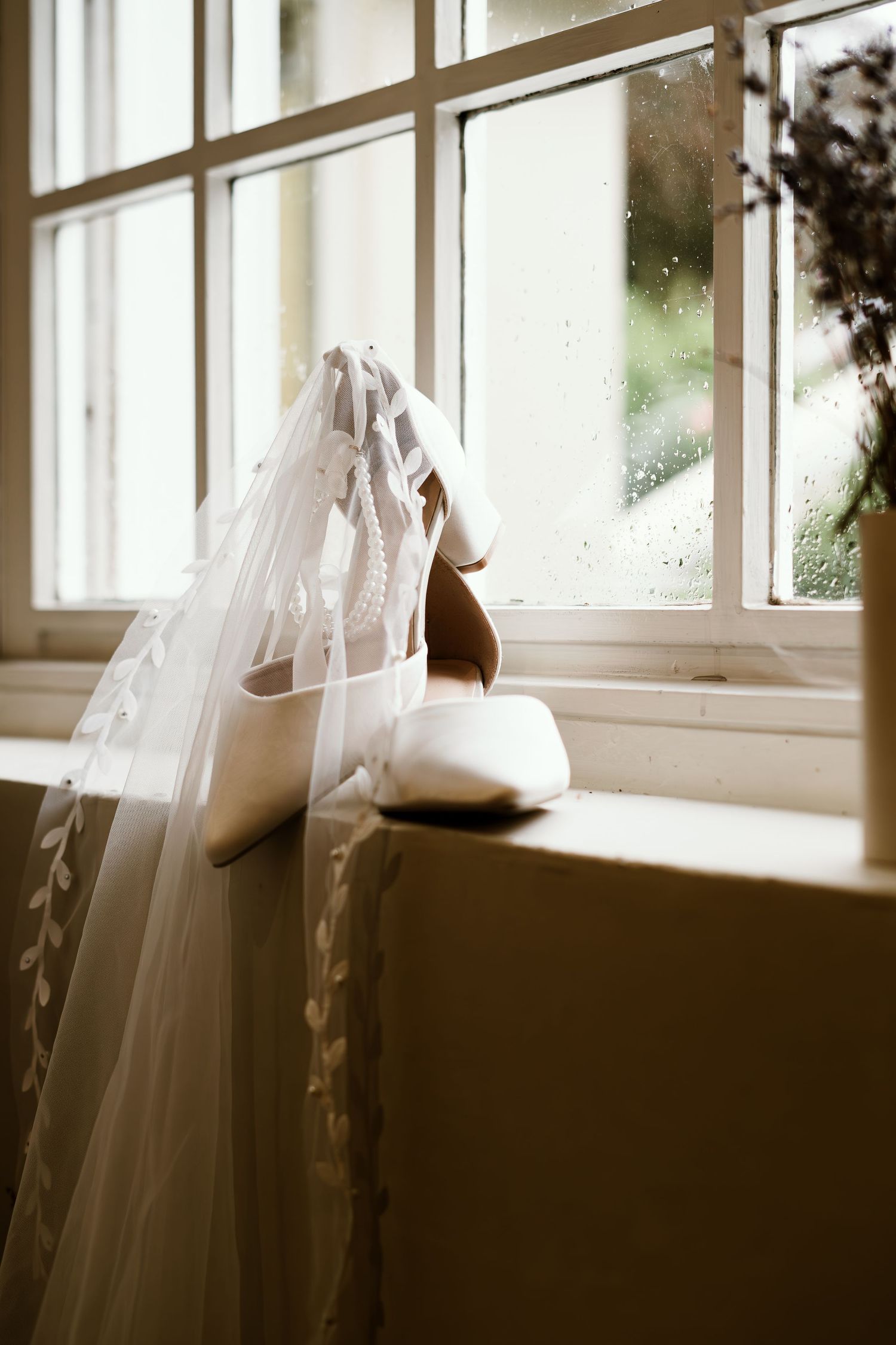 A delicate white wedding veil and shoes sit on a windowsill in soft natural light.