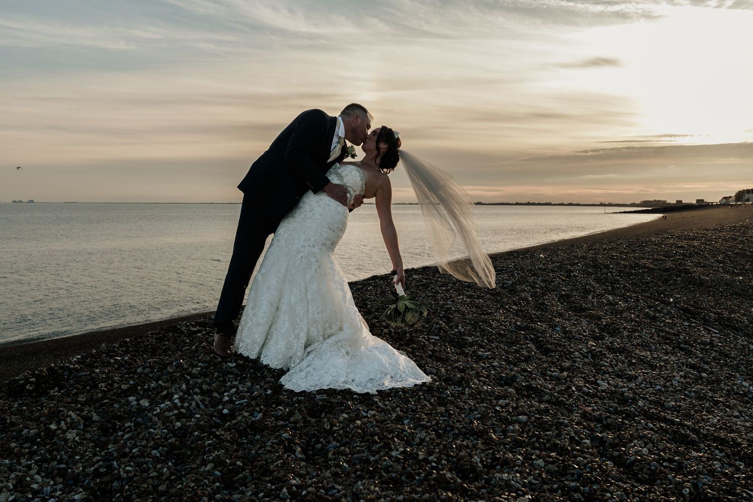 A romantic sunset beach wedding photoshoot with a couple embracing on a rocky shoreline.