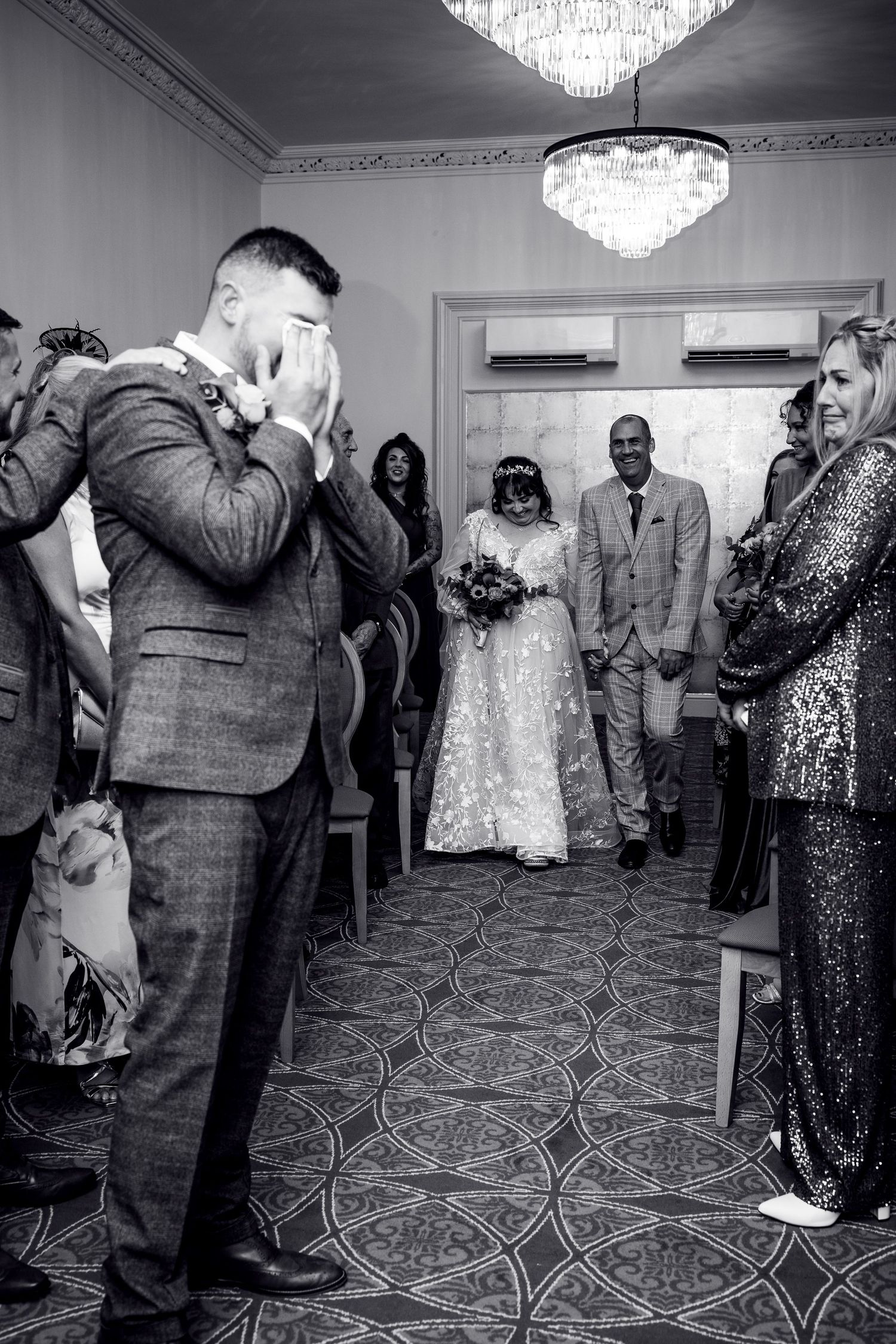 An emotional black and white wedding ceremony moment captured under crystal chandeliers.