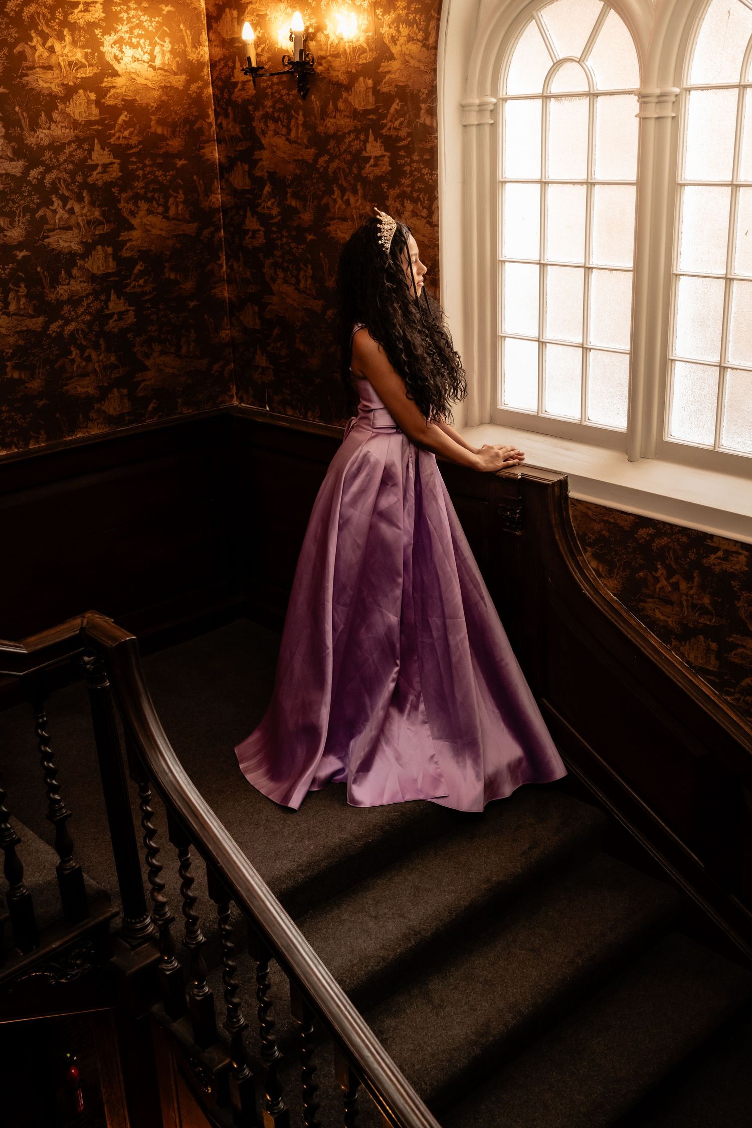 A person in a flowing purple ball gown stands elegantly on a grand staircase near an arched window.