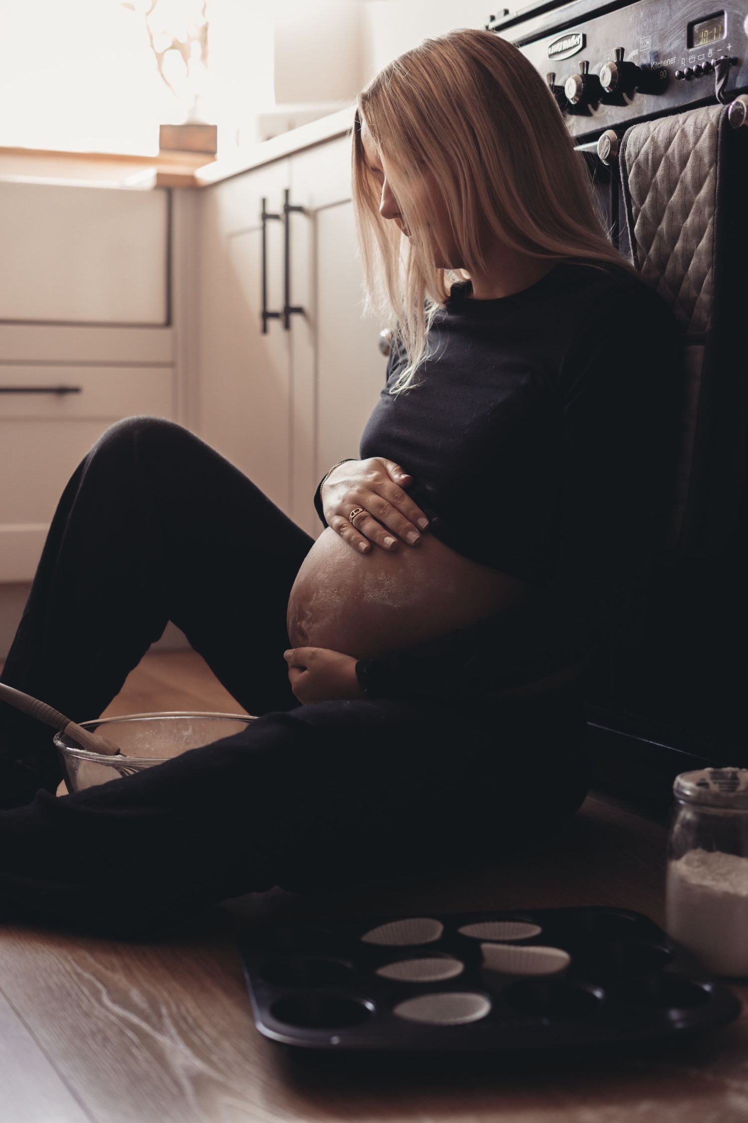 Pregnant woman in black clothing sits on kitchen floor cradling her belly.