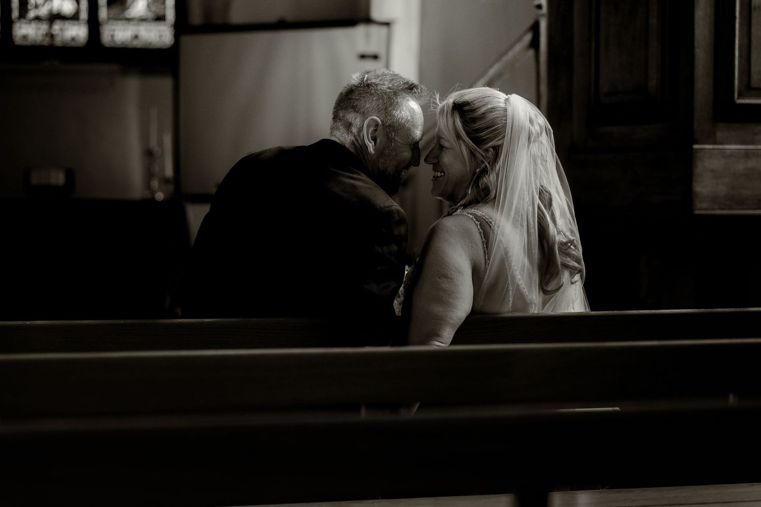 Intimate black and white photo of a couple sharing a moment in church pews.