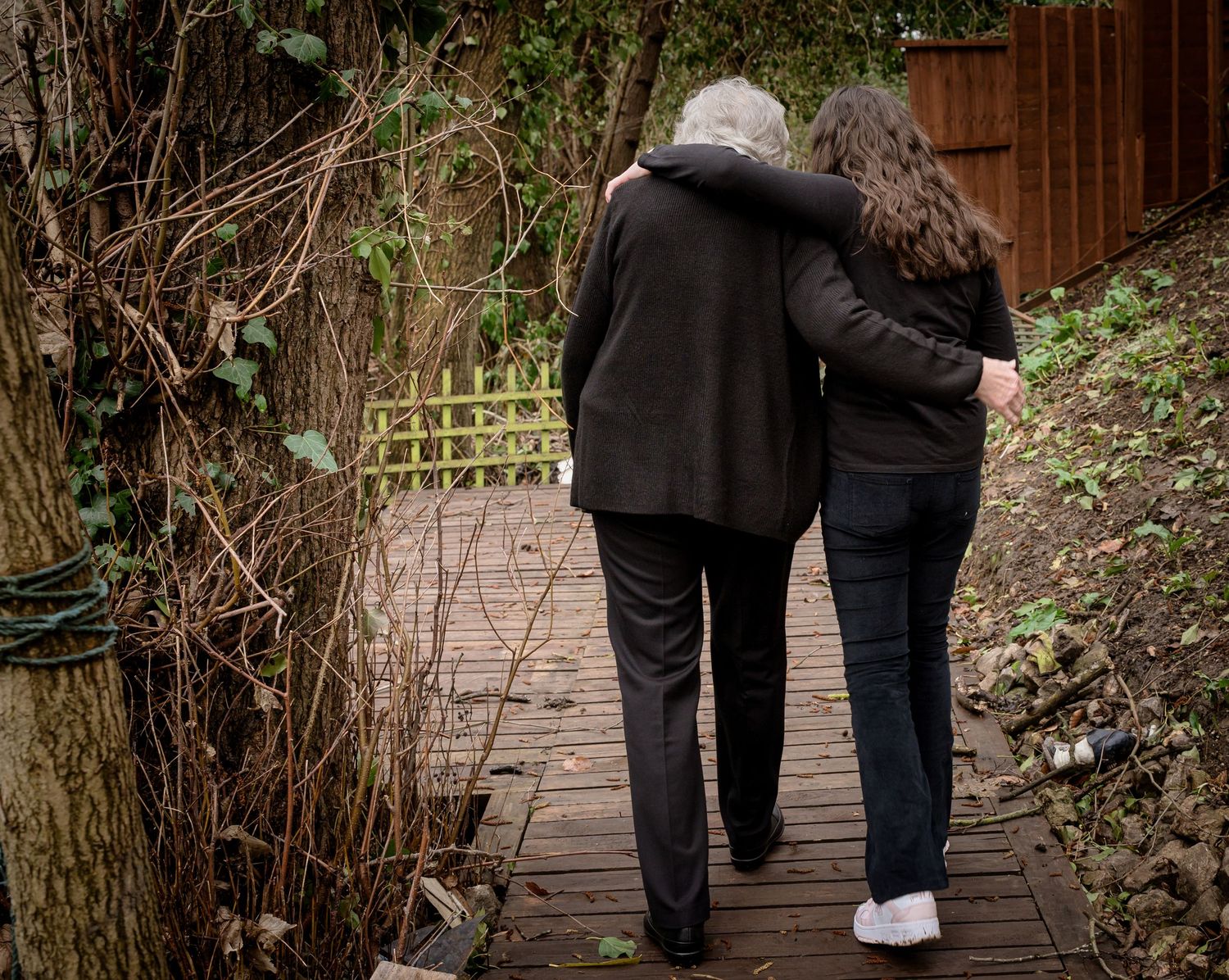 Two people walking together on a wooden pathway surrounded by winter foliage.