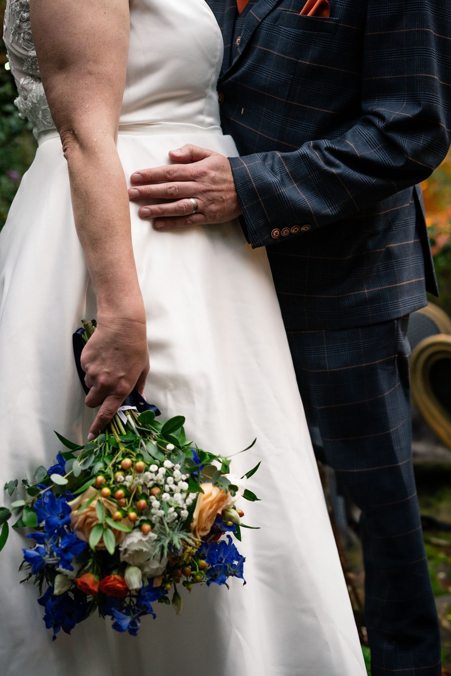 A bridal bouquet with blue flowers is held against a white wedding dress and navy pinstriped suit.
