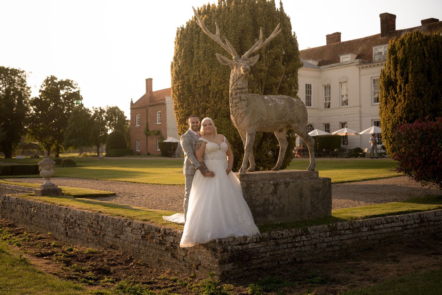 A wedding couple poses next to a large decorative deer statue in a stately garden setting at sunset.