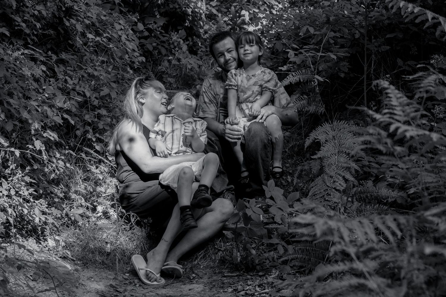 Family sitting together on forest steps surrounded by ferns in black and white.