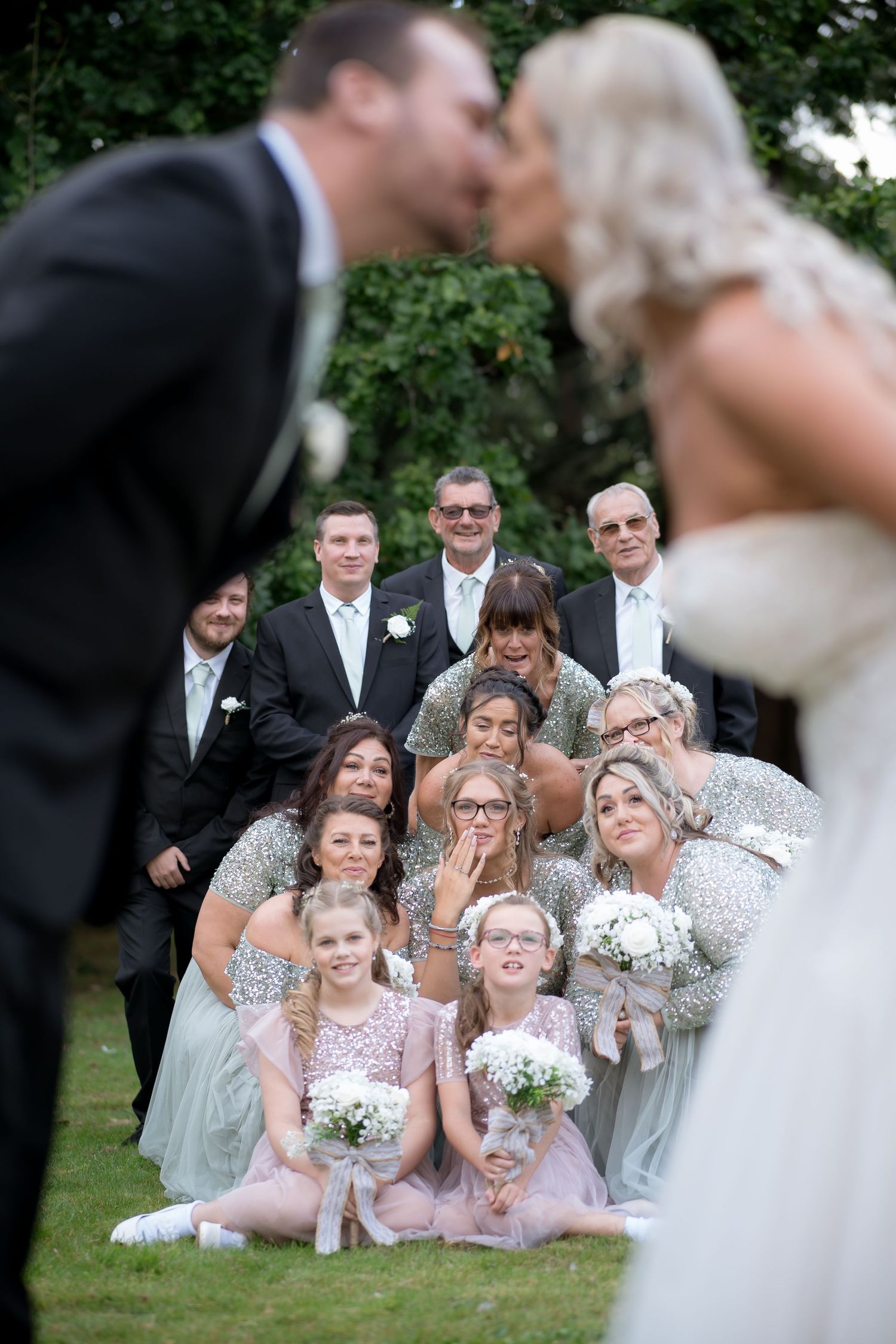 Creative wedding photo with bridal party reacting to couple's kiss in garden setting.
