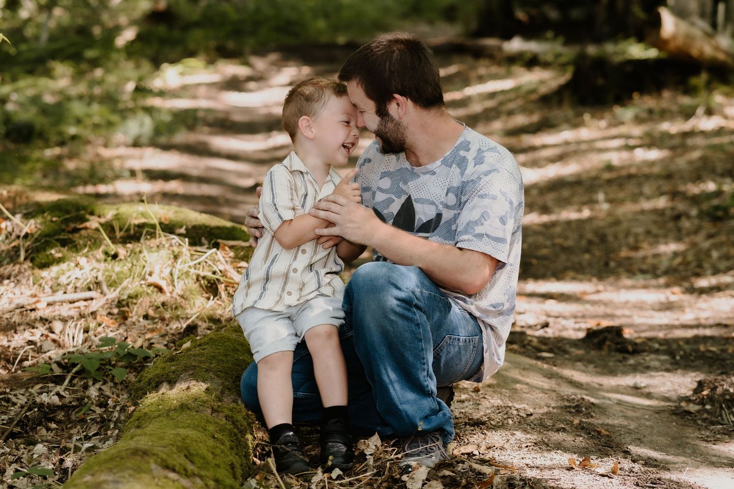 Two people share a tender moment while sitting on a mossy log in a sunlit forest setting.