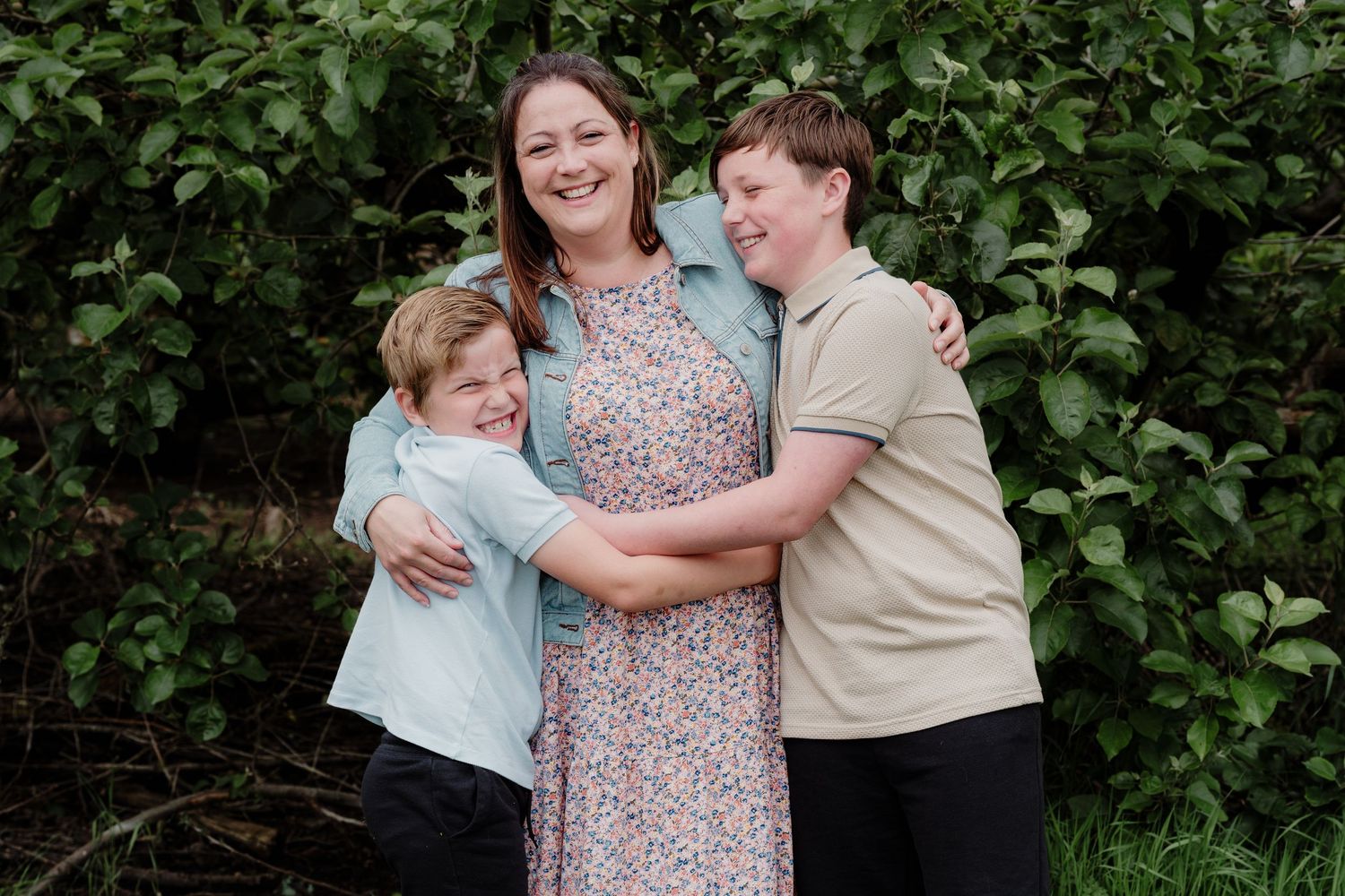 Three people share a warm embrace while standing in front of green bushes.