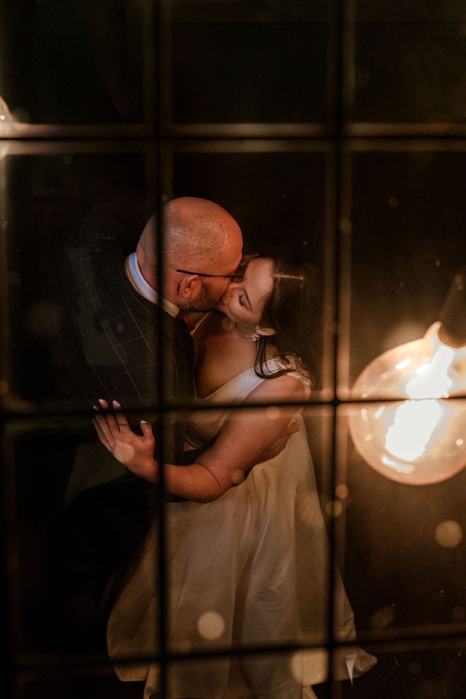 Romantic backlit wedding couple embrace through windowpane with glowing light fixture.