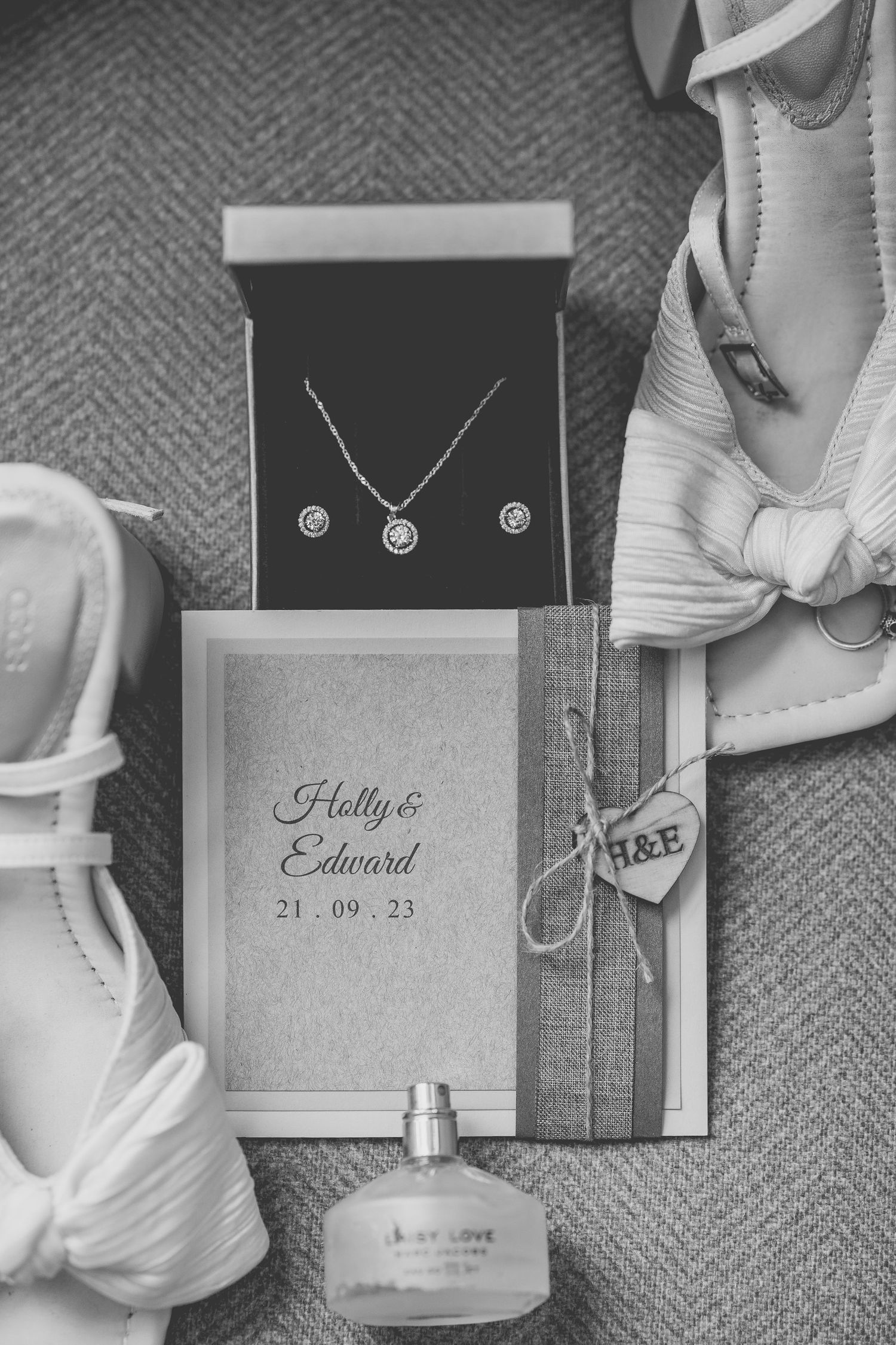 Wedding detail flatlay with jewelry box, white shoes, perfume bottle and wedding invitation on gray fabric.