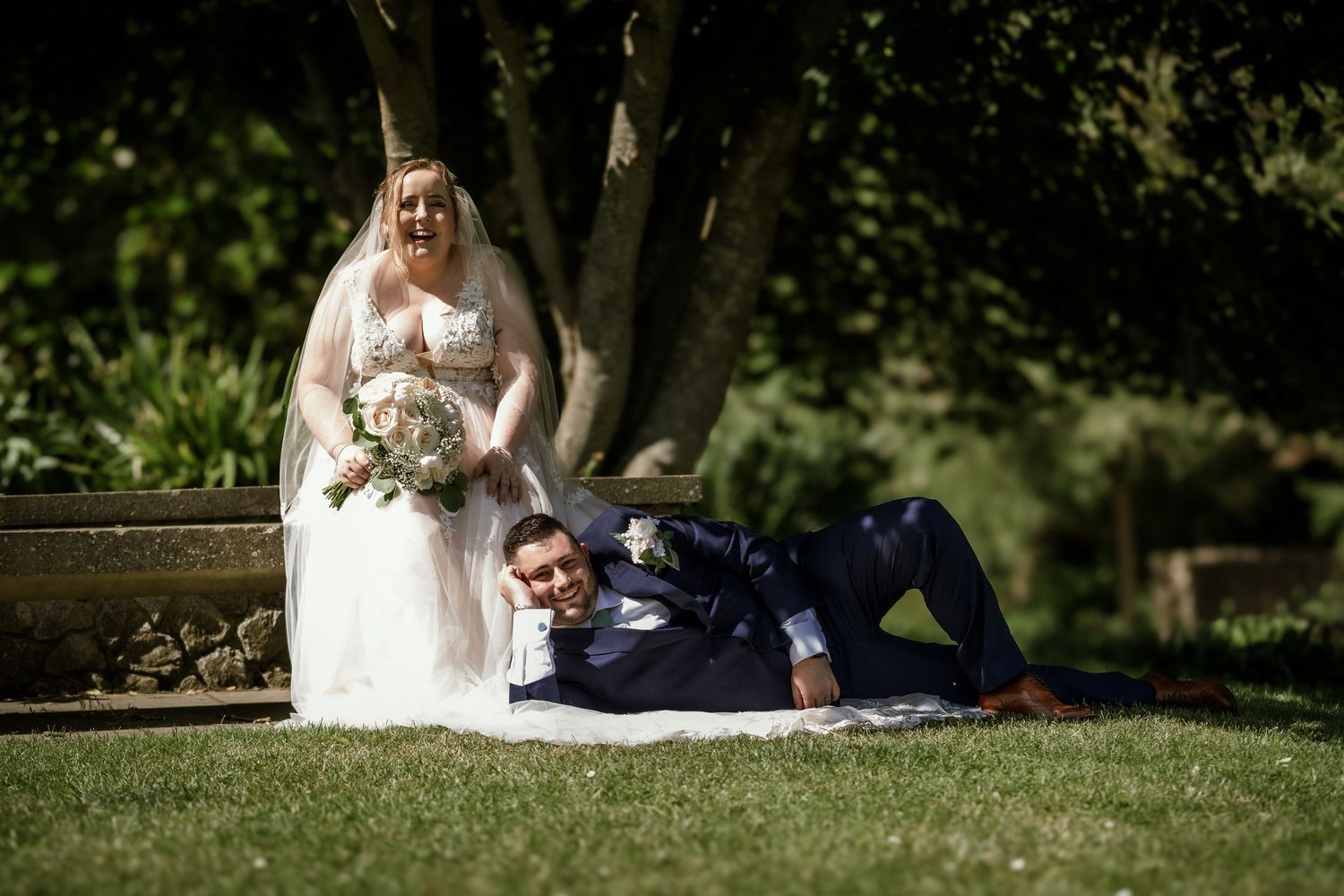 Playful wedding photo with bride sitting on bench and groom lying on grass.