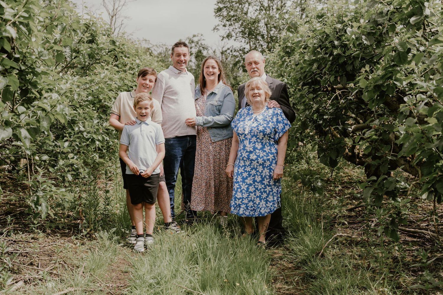Family of five poses together outdoors in casual attire against a backdrop of green foliage.