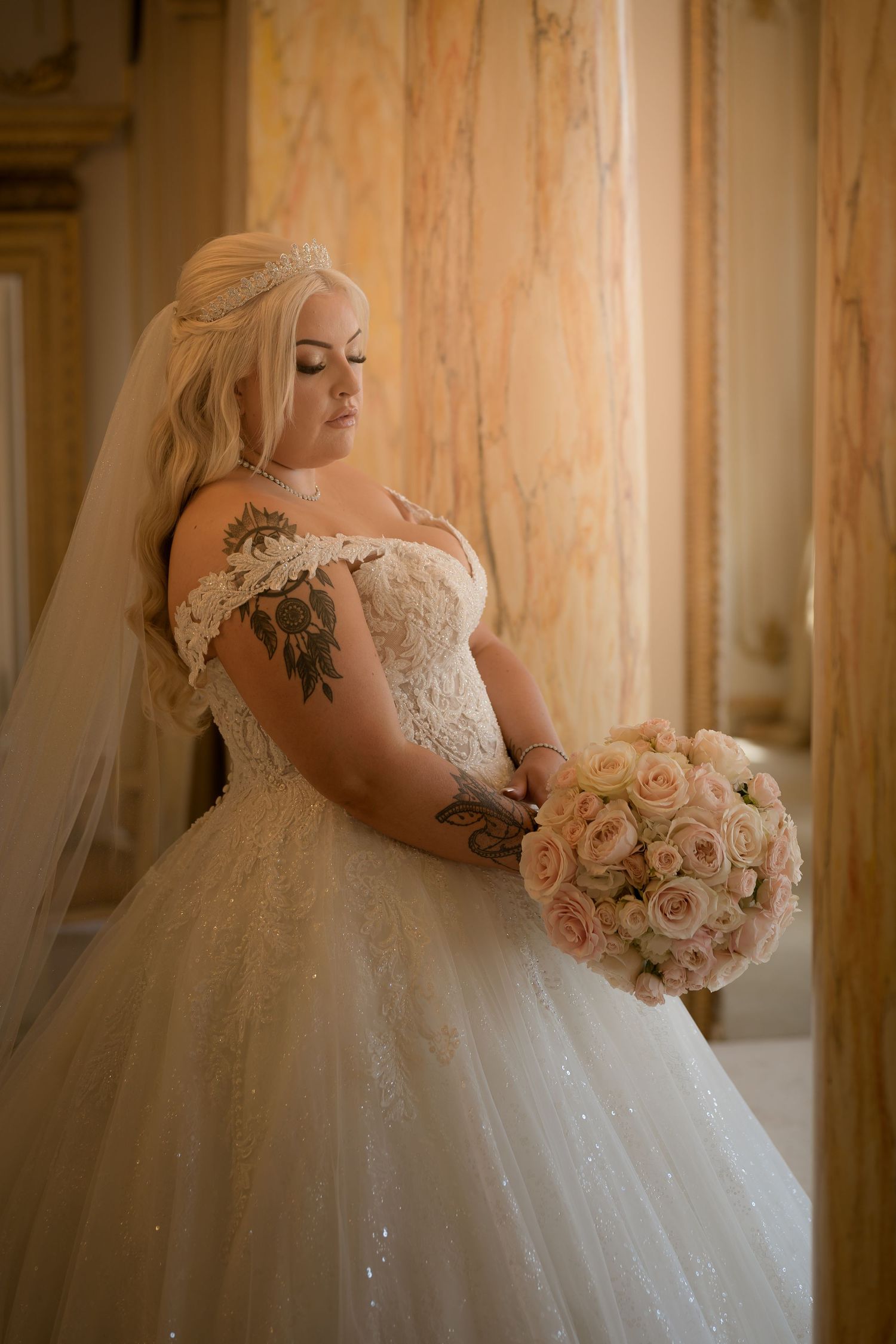 A bride in an off-shoulder lace gown holds a pink bouquet in an elegant setting.