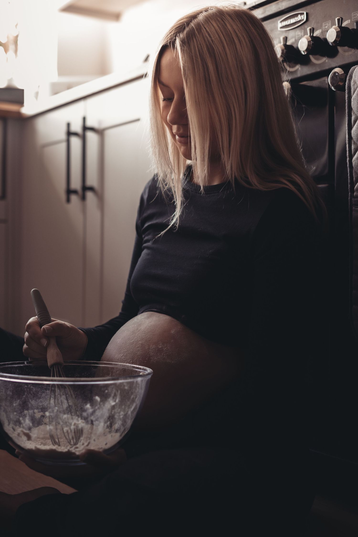 Pregnant woman in dark clothing mixing ingredients in bowl while sitting on kitchen floor.