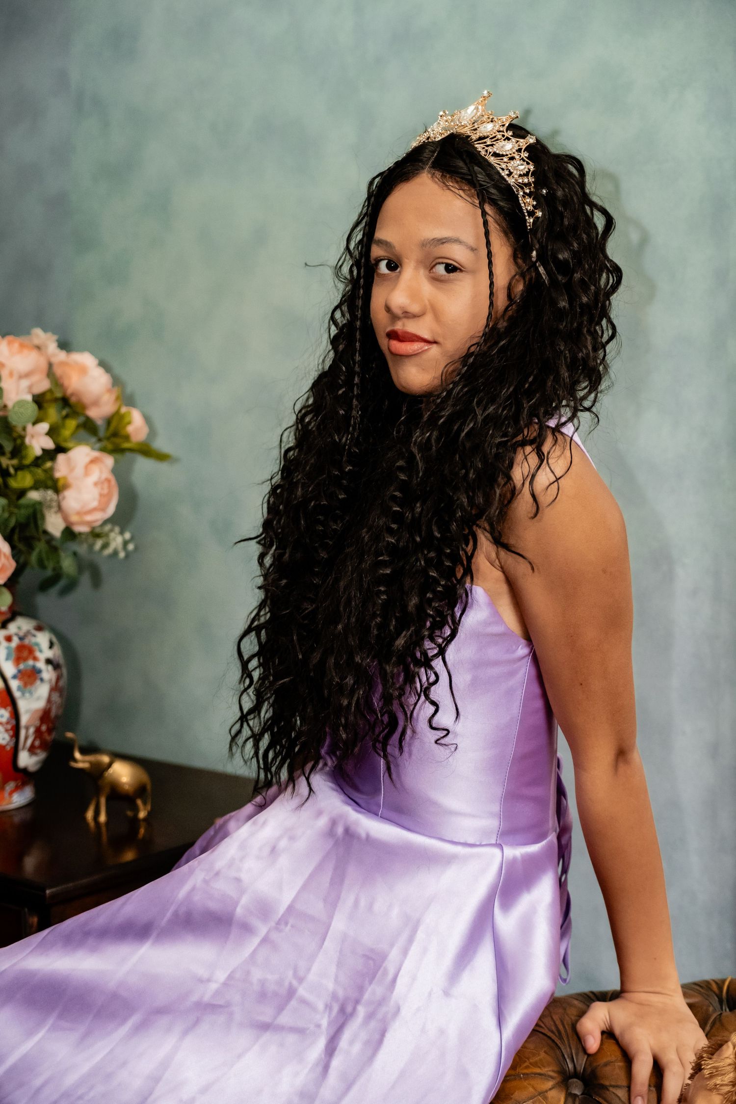 A person in a lavender gown and crown poses beside floral arrangements in a studio setting.