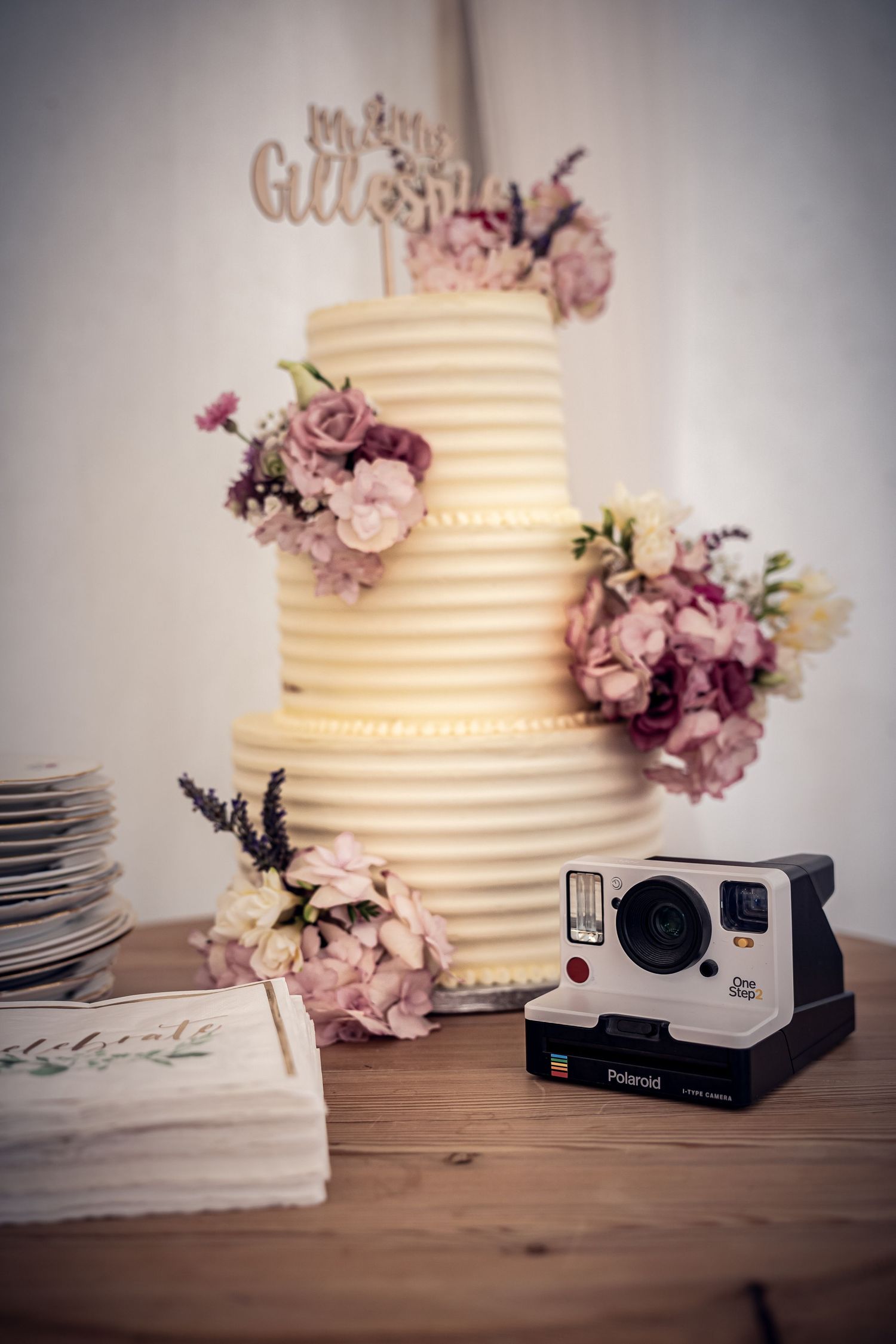 An elegant three-tier wedding cake with pink floral decorations next to a Polaroid camera.