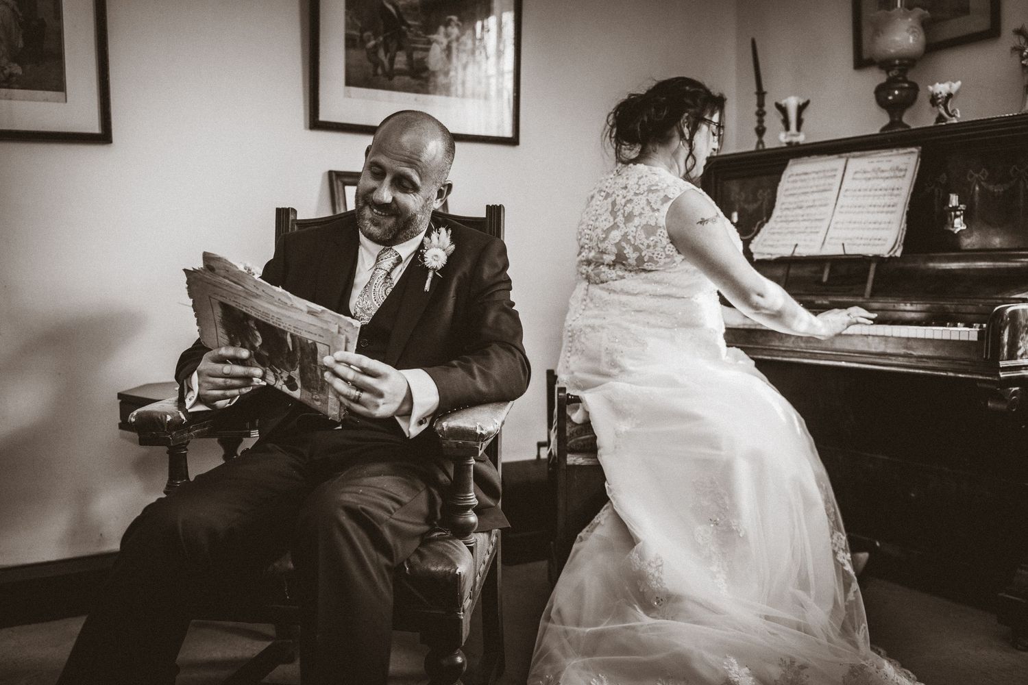 A candid black and white photo of a couple relaxing by a piano during their wedding day.