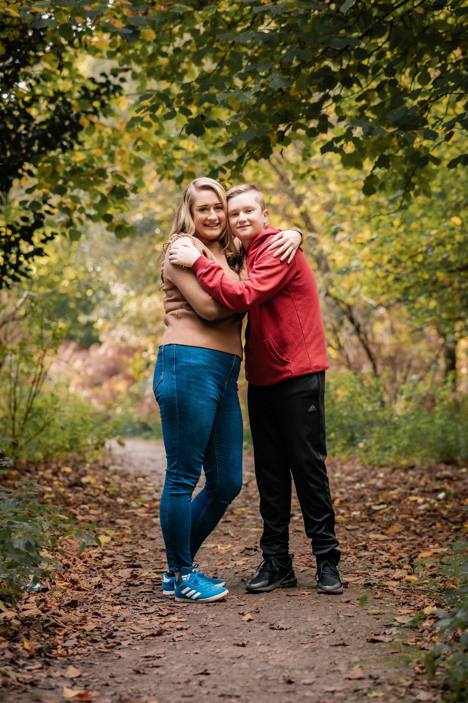 Two people embrace while standing on a leaf-covered autumn trail surrounded by trees.
