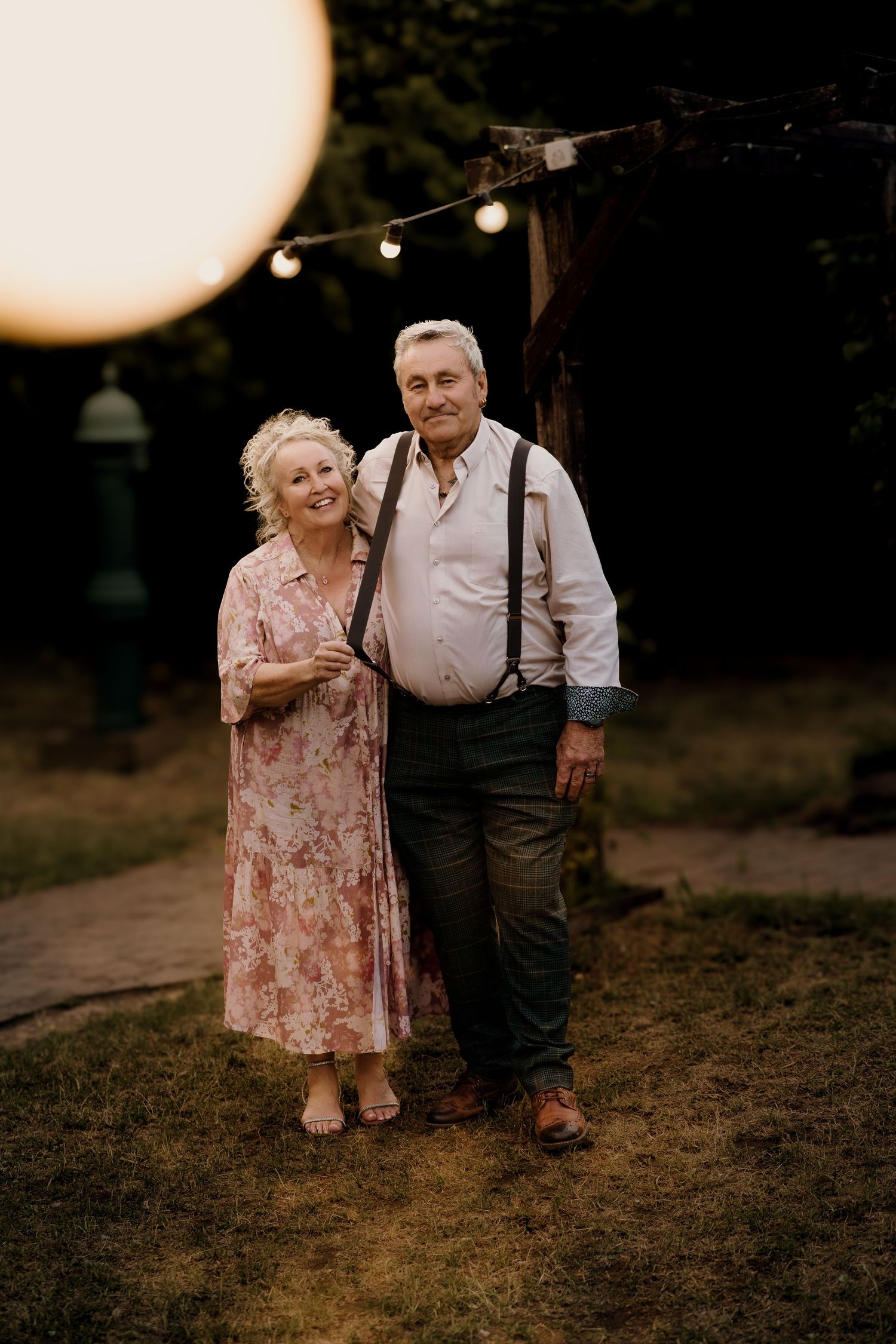 An elderly couple stands together outdoors at dusk under string lights, wearing a floral dress and suspenders.