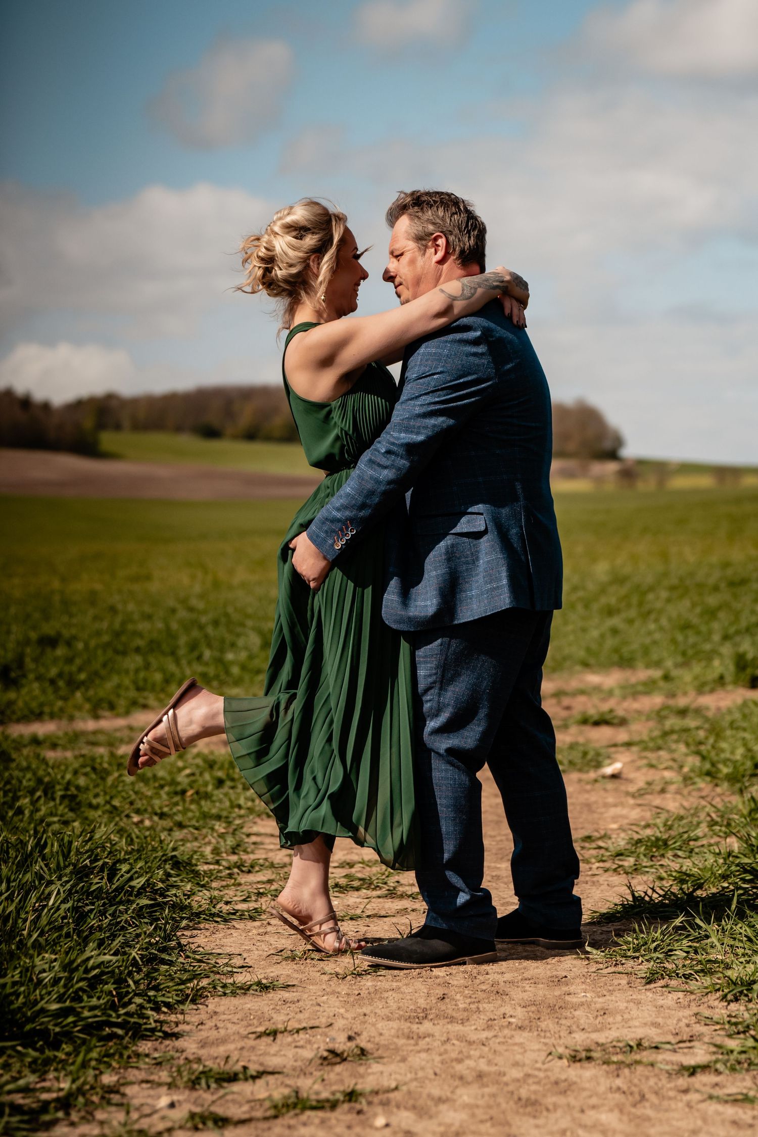 Romantic couple embraces while sitting on a dirt path through green fields on a cloudy afternoon.