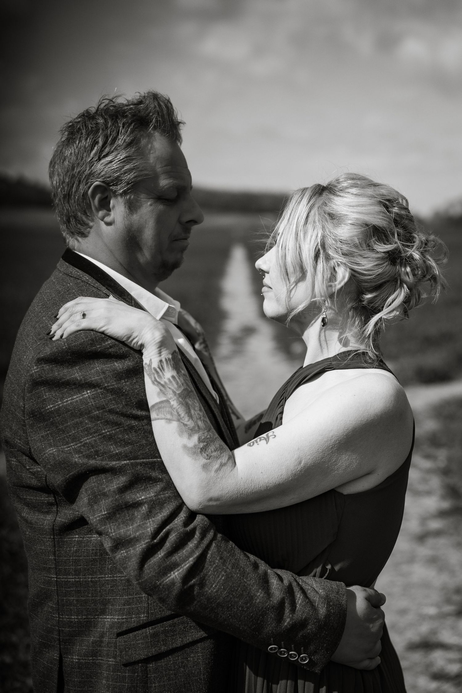 A black and white romantic portrait of a couple embracing outdoors, both dressed formally in vintage style clothing.