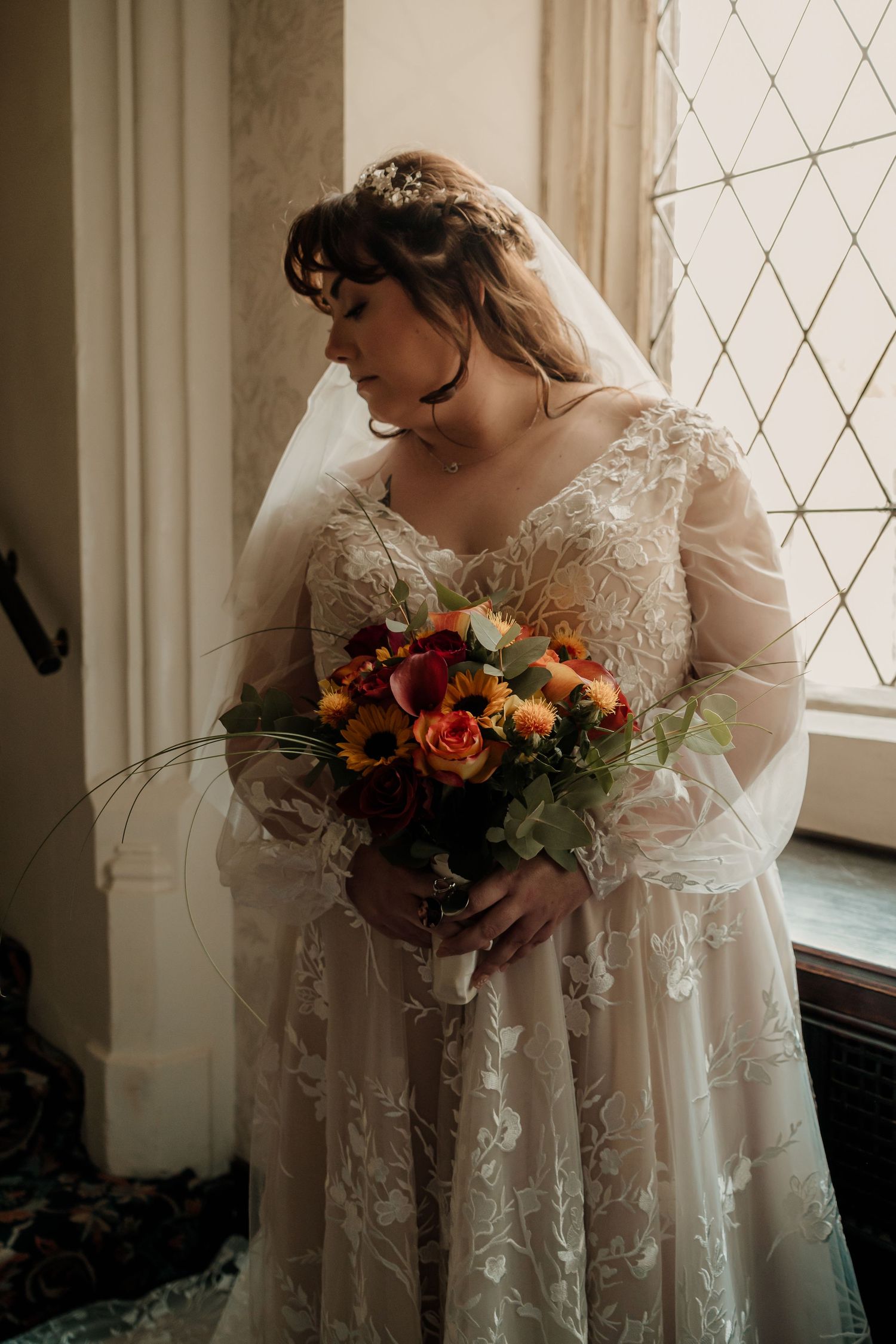 Bride in long-sleeve lace wedding dress holds vibrant fall-colored bouquet while standing by latticed window.