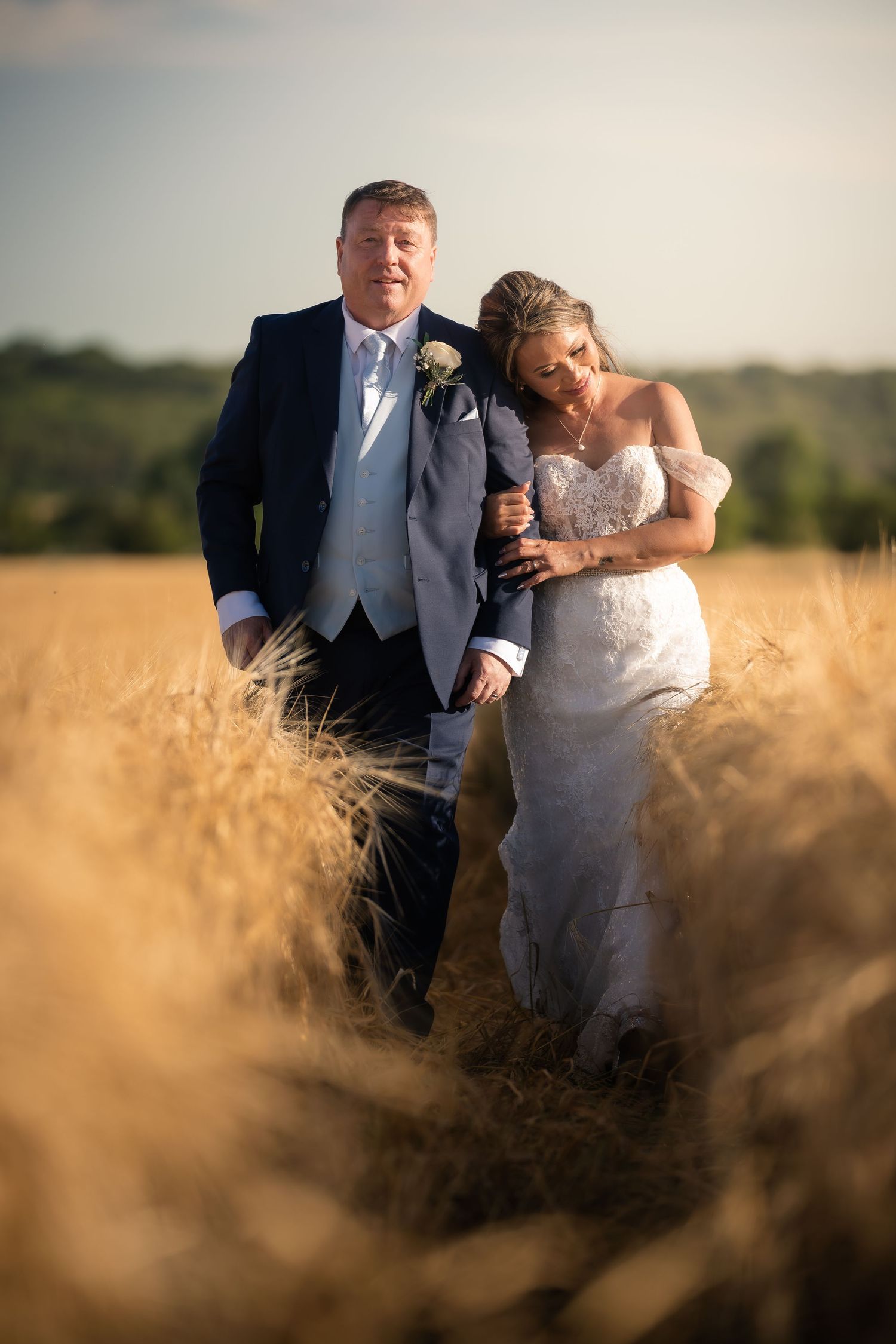 Wedding couple stands together in tall golden wheat field during sunset.
