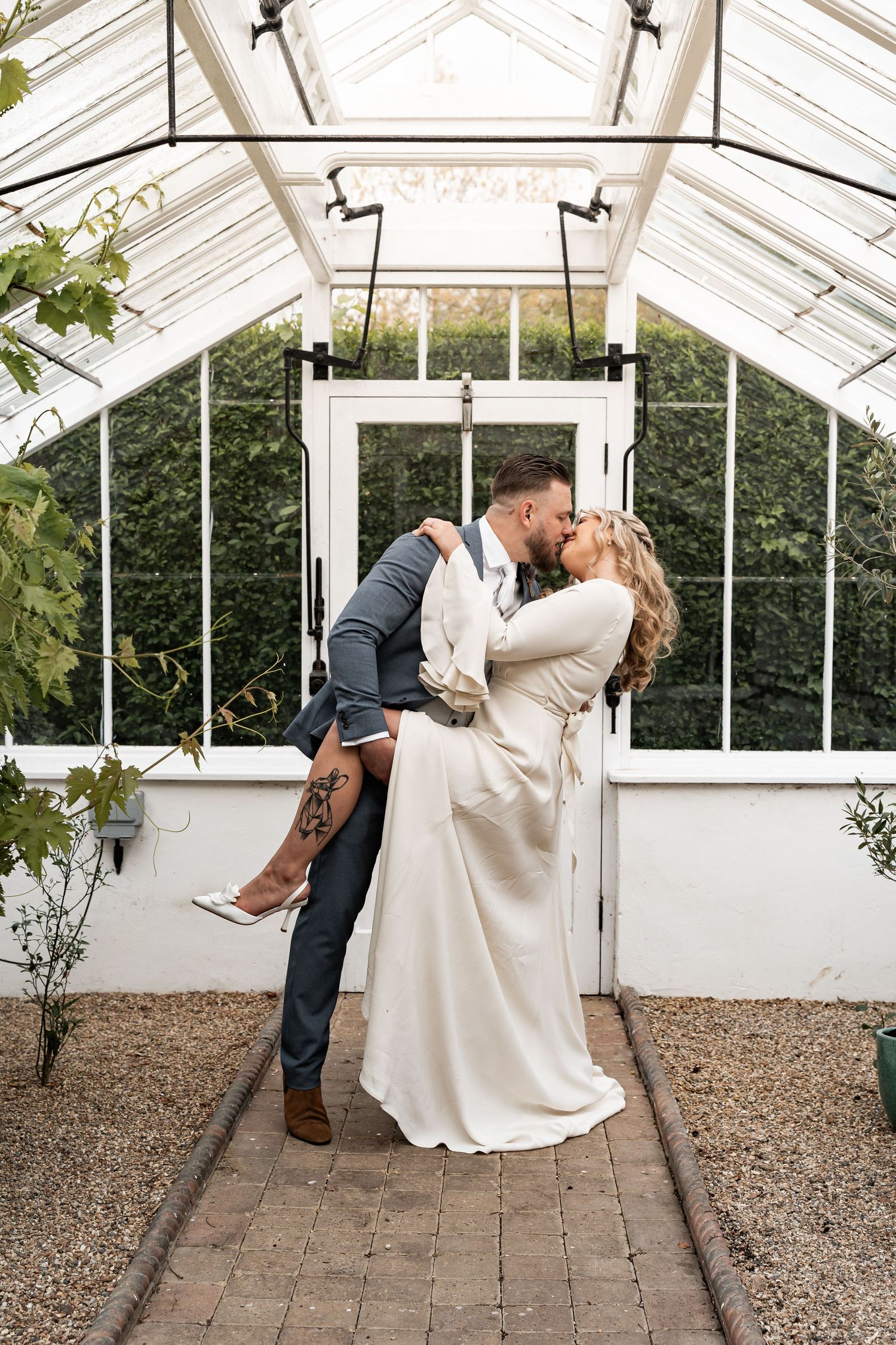 Romantic wedding couple embracing in a sunlit greenhouse with glass walls and brick path.