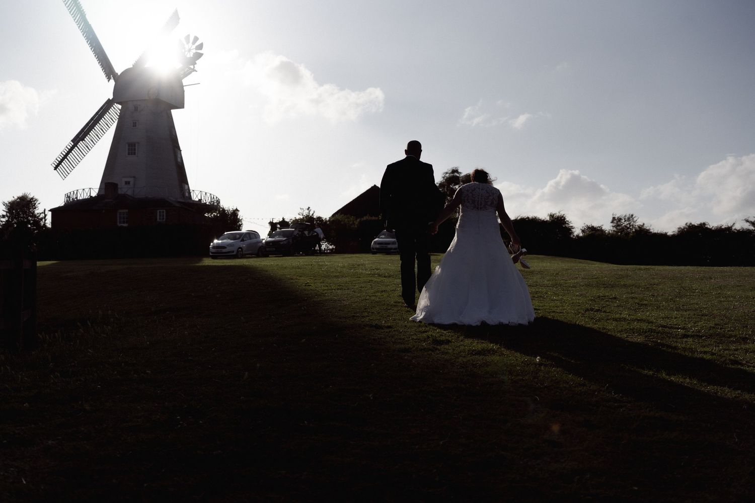 A couple walking towards a windmill silhouetted against a bright sky on their wedding day.