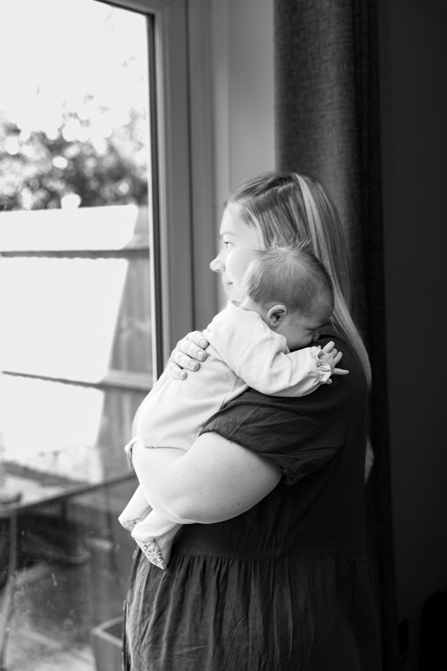 Black and white image of a woman holding a baby while looking out a window.
