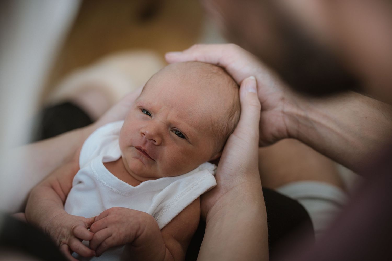 A newborn baby in a white onesie rests while being cradled in gentle hands.