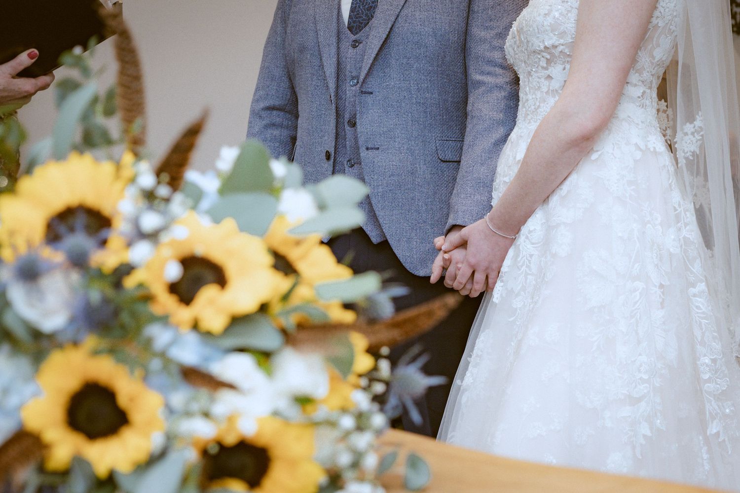 Wedding ceremony detail showing couple holding hands with sunflower bouquet in foreground.