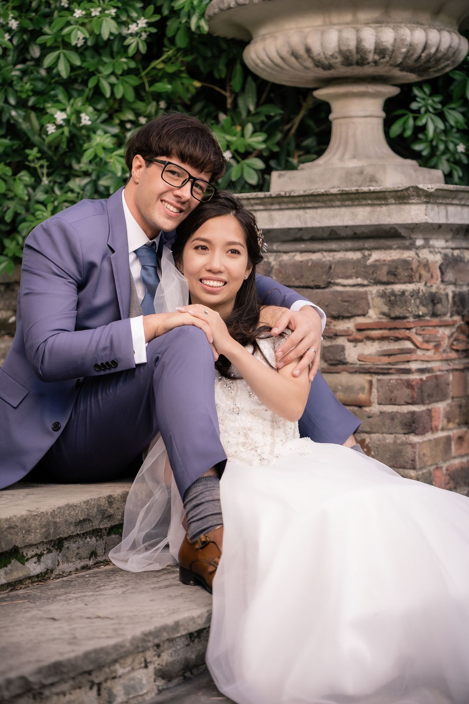 A couple in wedding attire poses together near an ornate garden urn.