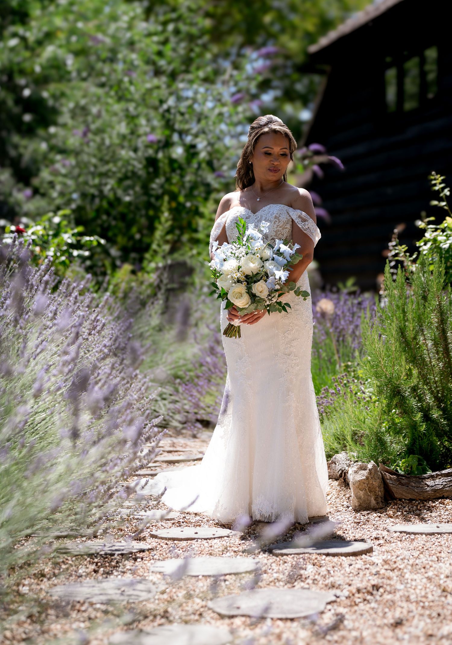 A bride in a white lace gown poses in a garden path surrounded by lavender and greenery.