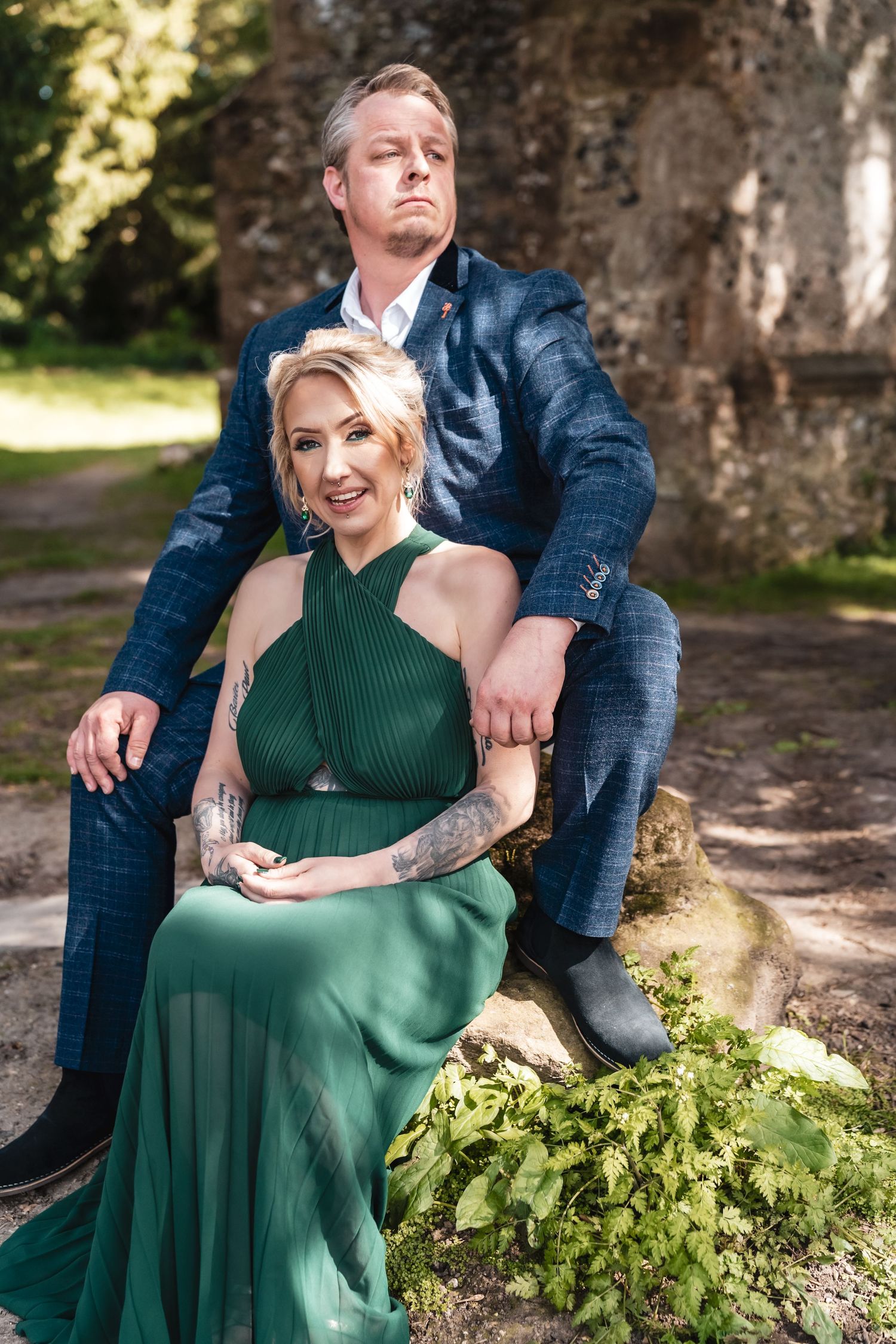 A couple poses together outdoors, with her in an elegant emerald green dress and him in a navy checkered suit.