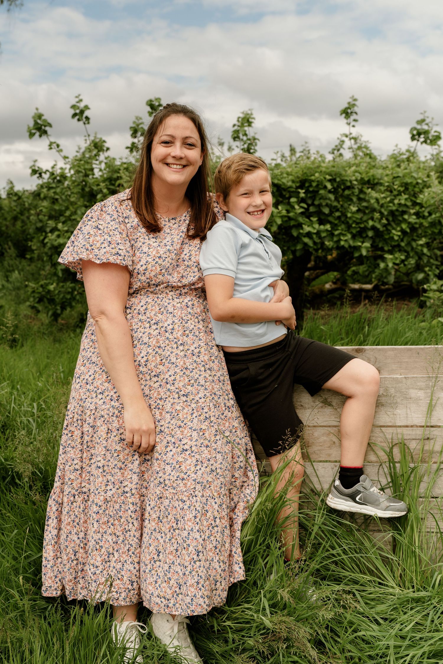 Two people pose together in a casual outdoor setting with green foliage and grass.