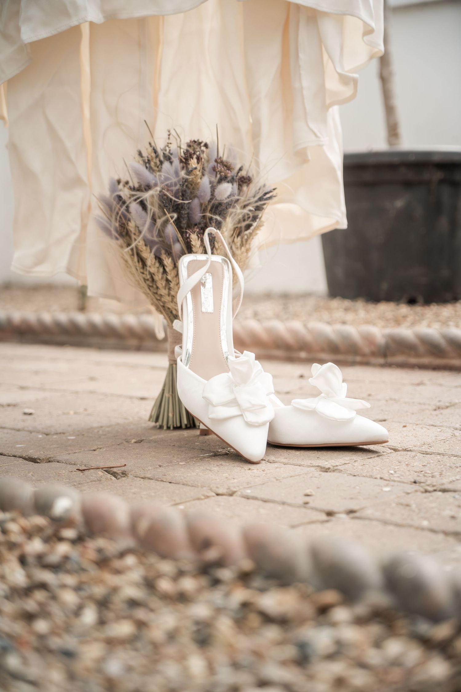 Elegant white bridal shoes placed next to a dried lavender bouquet on stone pavement.