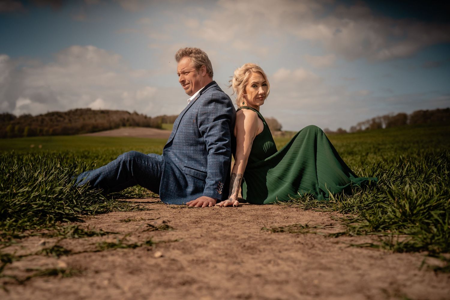 A couple sits together on a dirt path surrounded by green fields on a sunny day creating romantic silhouettes.